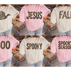 May include: A collection of six short-sleeved t-shirts in cream and light pink. Designs include a flying duck, and text graphics reading JESUS, FALL, BOO, SPOOKY, and SPOOKY SEASON, featuring various animal print and camouflage patterns.