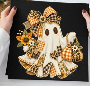 Op de afbeelding: Een vierkant zwart canvas met een centrale spookfiguur versierd met stoffen lapjes in oranje, bruin en luipaardprint. Het spook draagt een grote strik, houdt een pompoenmandje vast en is versierd met zonnebloemen en kleine witte bloemen.