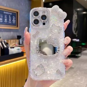 May include: A clear phone case with a white pearlescent finish and a sparkling Hello Kitty design. The case features a large Hello Kitty face cutout, a bow, and other decorative elements, all encrusted with rhinestones. The case is designed for a smartphone with multiple camera lenses.