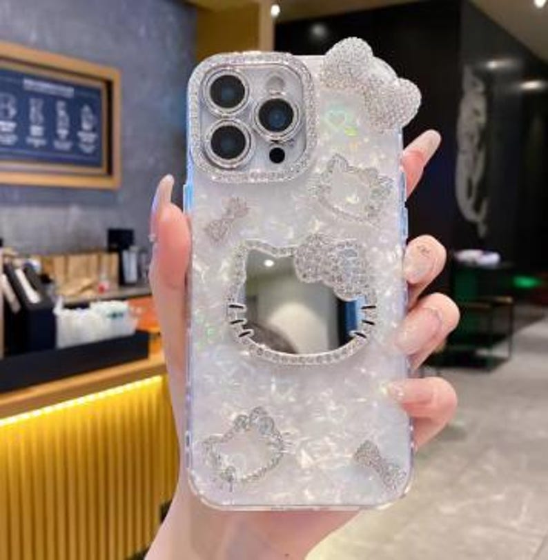 May include: A clear phone case with a white pearlescent finish and a sparkling Hello Kitty design. The case features a large Hello Kitty face cutout, a bow, and other decorative elements, all encrusted with rhinestones. The case is designed for a smartphone with multiple camera lenses.