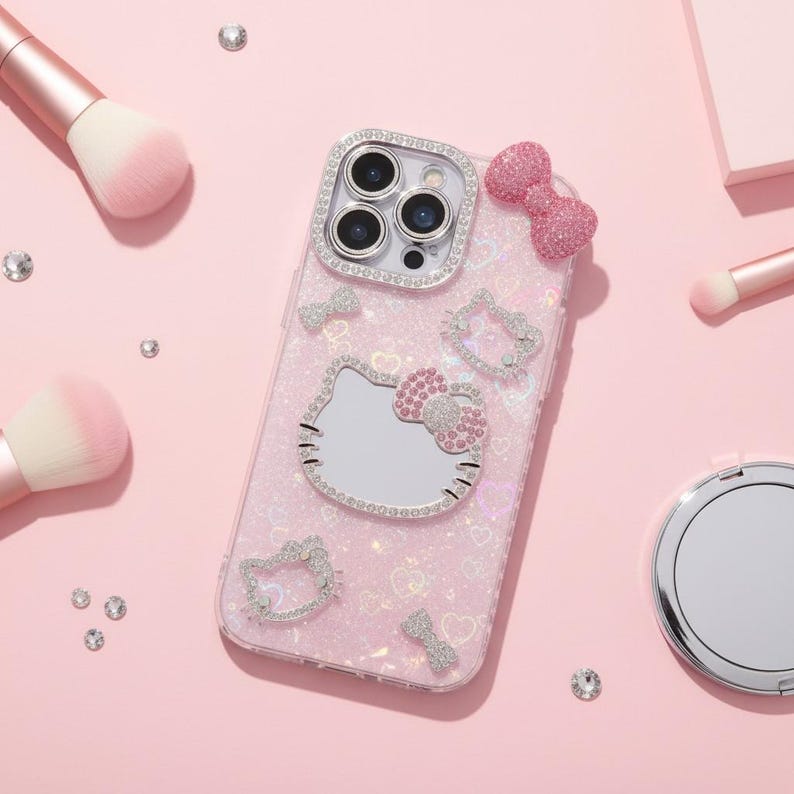 May include: A pink phone case featuring a Hello Kitty design, adorned with rhinestones. The case has a bow and several Hello Kitty outlines, with a mirrored face. Makeup brushes and loose rhinestones are scattered around the case on a pink surface.