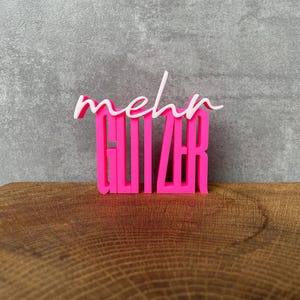 Könnte beinhalten: Ein leuchtend pinkfarbenes Schild mit der Aufschrift "mehr GLITZER". Das Wort "mehr" ist in weißer Schrift geschrieben, während "GLITZER" in einer kräftigen, pinkfarbenen Blockschrift gehalten ist. Das Schild steht auf einer Holzoberfläche.