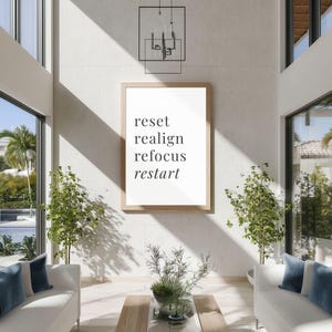 May include: A framed print with the words "reset, realign, refocus, restart" in a modern font. The print is displayed in a bright, airy living room with white sofas, blue pillows, and potted plants. A contemporary light fixture hangs above.