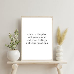 May include: A framed print with the text "stick to the plan not your mood not your feelings not your emotions." The print is displayed on a light wooden shelf with a white vase holding flowers on the left and a white vase with dried grass on the right.