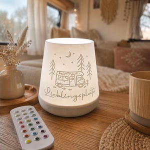 Könnte beinhalten: Eine weiße, konische, beleuchtete Lampe mit einer Strichzeichnung eines Wohnmobils, Tannenbäumen, einer Mondsichel und Sternen. Der Text "Lieblingsplatz" ist in Schreibschrift unter dem Wohnmobil aufgedruckt. Eine weiße Fernbedienung.