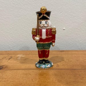 May include: A decorative Christmas nutcracker figurine. The figurine is dressed in a red jacket with gold trim, a black hat, and a white beard. It holds a gold sword and stands on a floral base. The nutcracker is approximately 15 cm tall.