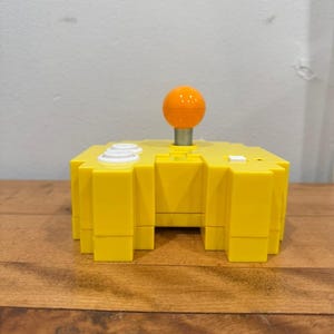 Pode incluir: Um joystick de estilo retrô amarelo brilhante com uma alça laranja em forma de bola. O controlador tem botões brancos e uma base quadrada. O dispositivo está em uma superfície de madeira.