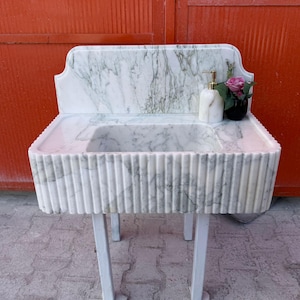 May include: A white marble sink with a fluted front design, set on a white metal stand. The sink features a rectangular basin and a backsplash. A marble soap dispenser and a small vase with pink flowers are on the countertop.