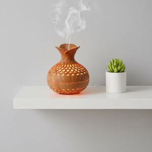 Peut inclure: Un diffuseur d'huiles essentielles en bois avec un dessus en forme de fleur et un corps à motifs. Le diffuseur émet un flux de vapeur. Une petite plante succulente verte dans un pot blanc se trouve à droite du diffuseur.