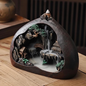 Peut inclure: Un brûleur d'encens en céramique marron en forme de scène de montagne. Le brûleur présente une cascade, des maisons miniatures, des arbres et un pont. Un cône d'encens blanc est placé au sommet, avec de la fumée s'élevant de la base, créant un effet brumeux.