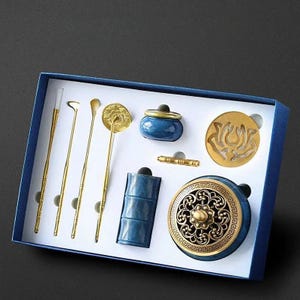 Peut inclure: Un ensemble de boîtes d'outils et d'accessoires d'encens dorés. L'ensemble comprend plusieurs outils fins, un petit pot en céramique bleu, un porte-encens bleu et un brûleur d'encens doré avec un motif de fleur de lotus.