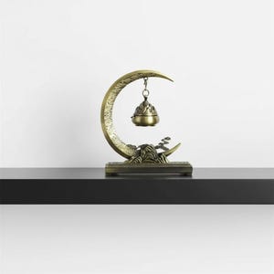 Peut inclure: Un brûleur d'encens de couleur bronze en forme de croissant de lune. Le brûleur présente un motif détaillé de montagne et d'arbre sur la base et un porte-encens suspendu. Le design est orné et décoratif.