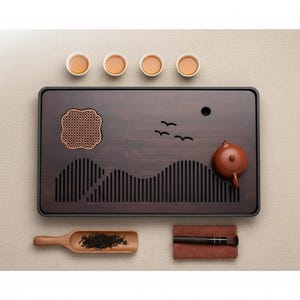 Peut inclure: Plateau à thé rectangulaire marron foncé avec bordure noire, orné d'un motif montagne sculpté et d'une petite théière. Quatre petites tasses à thé blanches avec du thé sont disposées au-dessus du plateau. Une cuillère et une brosse en bois sont également incluses.