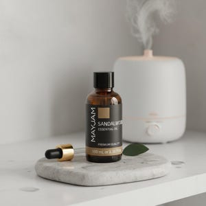 Peut inclure: Une bouteille en verre brun d'huile essentielle de bois de santal MAYJAM, 100 ml, avec une étiquette et un bouchon noirs. Un compte-gouttes or et noir et un diffuseur blanc sont également visibles. La bouteille est posée sur un plateau en marbre gris avec une feuille verte.