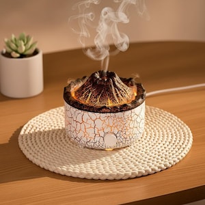 Peut inclure: Diffuseur d'huiles essentielles en forme de volcan blanc et marron avec un motif craquelé. Le sommet du diffuseur est de couleur marron foncé et émet un flux de vapeur. Il repose sur un tapis rond en tissu crème.
