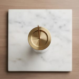 Peut inclure: Un porte-encens en laiton avec un bâton d'encens marron, posé sur un carré de marbre blanc. Le support a une base cylindrique et un dessus circulaire. Le marbre présente des veines grises, et le fond est en bois brun clair.
