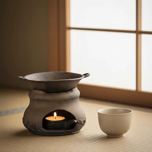 Peut inclure: Un brûleur d'huile en céramique avec une petite bougie allumée à l'intérieur, à côté d'une petite tasse à thé blanc cassé. Le brûleur a un plat gris foncé sur le dessus et des motifs floraux décoratifs. La scène est devant une fenêtre.