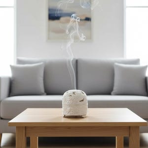 Peut inclure: Un brûleur d'encens en céramique blanche avec un motif floral, dégageant de la fumée. Le brûleur est posé sur une table en bois brun clair, devant un canapé et des coussins gris. La pièce a une palette de couleurs neutres.