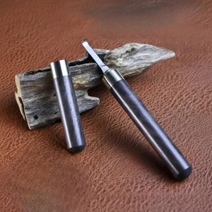 Puede incluir: Un juego de herramientas para tallar madera con un mango de madera oscura y una cuchilla de metal plateado. El juego incluye una cuchilla y una funda protectora. Las herramientas se muestran sobre una superficie marrón.