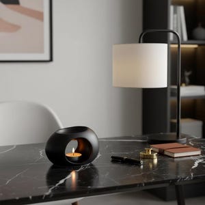 Peut inclure: Un diffuseur d'huiles essentielles noir de forme ovale avec une bougie allumée à l'intérieur, posé sur une surface en marbre noir. Un abat-jour blanc et une lampe de bureau noire sont en arrière-plan. Un stylo, un petit carnet et un objet doré sont également sur la table.