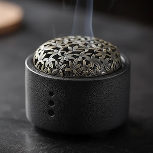 Peut inclure: Un brûleur d'encens en céramique noire avec un couvercle décoratif de couleur bronze orné d'un motif floral. De la fumée s'élève doucement du dessus. Le brûleur est de forme cylindrique avec trois petits trous sur le côté.