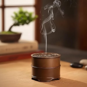 Peut inclure: Un brûleur d'encens en métal marron de forme cylindrique avec un dessus décoratif. Des volutes de fumée blanche s'élèvent du brûleur, suggérant de l'encens en combustion. Un petit bonsaï est visible en arrière-plan.
