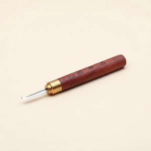 Può includere: Uno strumento per intaglio con manico in legno e lama curva. Il manico è di colore marrone rossiccio scuro con dettagli metallici dorati. La lama è argentata. Lo strumento sembra nuovo.