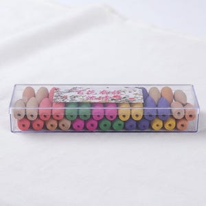 Peut inclure: Une boîte en plastique transparente remplie de cônes d'encens colorés. Les cônes sont disposés en rangées et sont de différentes couleurs, notamment rose, vert, jaune et violet. Une étiquette décorative avec des motifs floraux est visible.