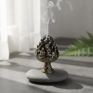 Peut inclure: Un brûleur d'encens orné de couleur bronze, dégageant de la fumée blanche. Le brûleur, au design complexe avec des motifs de flammes, est posé sur une pierre grise lisse. L'objet suggère une ambiance de relaxation et d'aromathérapie.