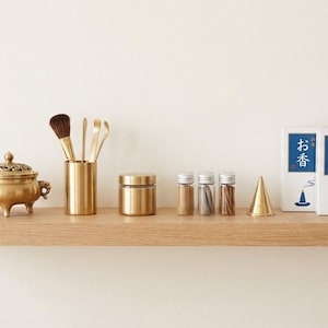 Peut inclure: Une collection d'objets dorés sur une étagère en bois. Comprend un brûleur d'encens décoratif, un pinceau, de petits pots, un cône et deux boîtes avec du texte et des illustrations japonais. L'étagère est contre un mur blanc uni.
