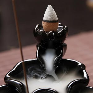 Puede incluir: Un quemador de incienso de cerámica negra con un palito de incienso en forma de cono. El incienso se está quemando, produciendo un humo blanco que cae en cascada por el quemador. El quemador tiene un acabado brillante y un diseño floral.