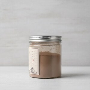 Peut inclure: Un pot en verre transparent avec un couvercle argenté, rempli d'une poudre brune. Une étiquette rectangulaire avec du texte et une illustration florale est apposée sur le pot. Le pot est posé sur une surface blanche.