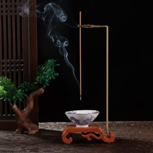 May include: An incense burner set with a gold-colored stand holding a lit incense stick. A small white bowl with blue floral designs sits on a wooden stand. Wisps of smoke rise from the incense stick against a dark background.
