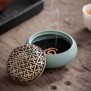 Peut inclure: Un brûleur d'encens en céramique bleu clair avec un couvercle décoratif doré orné d'un motif géométrique. De l'encens brûle à l'intérieur, dégageant de la fumée. Le brûleur est posé sur une surface en bois.