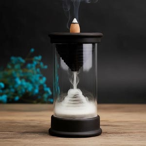 Peut inclure: Un brûleur d'encens en verre noir et transparent avec un bâton d'encens conique. Le brûleur a une base et un dessus noirs, avec un cône inversé noir à l'intérieur. De la fumée blanche s'élève du brûleur, créant un effet visuel.