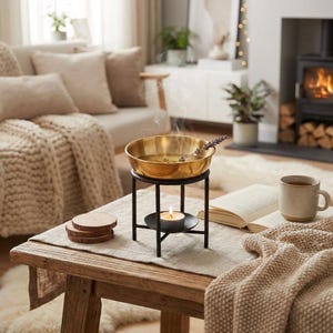Op de afbeelding: Aromatherapie oliebrander met een messing kom, een zwarte metalen standaard en een brandende kaars. De kom bevat water en een takje lavendel. De scène speelt zich af op een houten tafel met een neutrale loper, een boek en een mok.