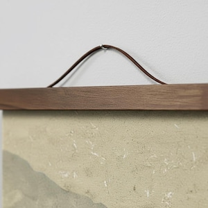 Peut inclure: Un support d'affiche en bois avec une corde en cuir marron pour l'accrocher. La barre en bois est de couleur marron moyen et maintient une œuvre d'art aux tons beige et gris.
