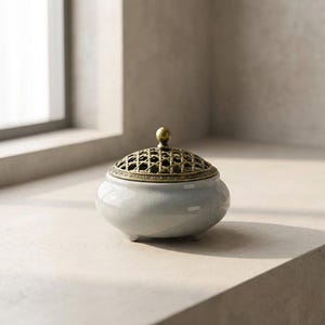 May include: A pale grey ceramic incense burner with a decorative brass lattice lid and a small brass knob. The burner has a rounded shape and sits on three small feet. The design is simple and elegant.