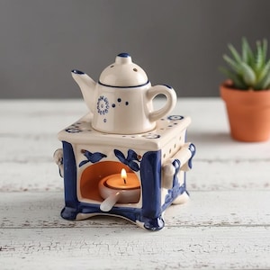 Peut inclure: Un chauffe-huile en céramique en forme de théière, avec un motif floral bleu et blanc. Le chauffe-huile a une petite théière sur le dessus et un porte-bougie à l'intérieur. Le design comprend des accents floraux bleus et une poignée. Une bougie allumée diffuse une lueur chaleureuse.