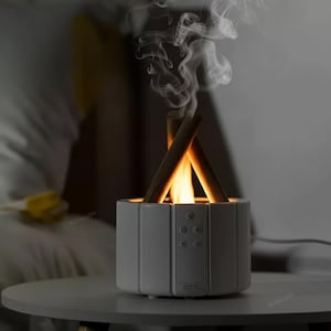 Peut inclure: Un diffuseur d'aromathérapie blanc, de forme hexagonale, avec un effet de flamme. Il présente des bâtons de bois disposés comme un feu de camp, avec des flammes et de la fumée simulées. Des boutons de commande sont visibles à l'avant.