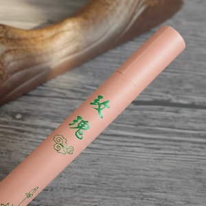 Peut inclure: Un tube cylindrique en papier rose clair avec des caractères chinois verts et des motifs de nuages. Le tube est en papier mat, probablement un contenant pour un produit.