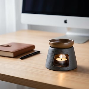 Peut inclure: Un diffuseur d'huiles essentielles en céramique noire avec un petit plat marron sur le dessus. Une bougie chauffe-plat allumée est à l'intérieur, dégageant une lueur chaleureuse. Sur une surface en bois clair, avec un carnet et un stylo.