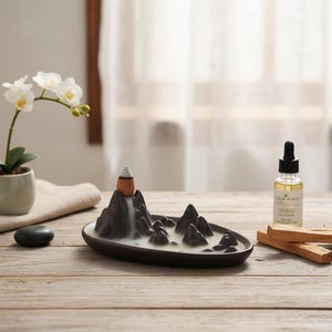 May include: A dark brown ceramic incense burner shaped like a mountain range, with a waterfall effect. A bottle of essential oil and palo santo sticks are next to the burner. A small potted orchid and a black stone are also in the image.