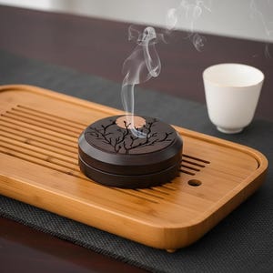 Peut inclure: Un brûleur d'encens brun foncé avec un motif d'arbre, dégageant des volutes de fumée. Il est posé sur un plateau en bambou, avec une tasse blanche en arrière-plan. Le couvercle du brûleur présente une silhouette d'arbre découpée.