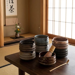 Peut inclure: Ensemble de bols à thé en céramique dans des tons bruns, gris et bleus, disposés sur une table en bois sombre. Un fouet et une cuillère en bambou sont également présents, suggérant une cérémonie du thé. Les bols ont un design texturé et strié.