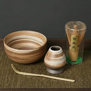 Peut inclure: Un ensemble à thé matcha comprenant un grand bol rayé marron et blanc, un petit vase, un fouet en bambou dans un récipient transparent et une cuillère en bambou. L'ensemble est pour préparer le thé matcha.
