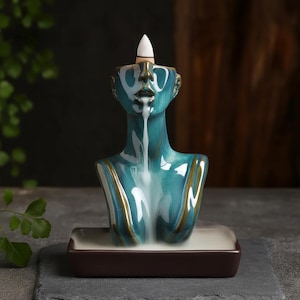 Peut inclure: Un brûleur d'encens en céramique turquoise en forme de buste, avec un bâton d'encens conique blanc. Le buste a une finition brillante avec des accents marron. La fumée s'échappe de la bouche, créant un effet visuel. Le brûleur repose sur une base rectangulaire marron.