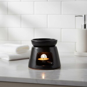 Peut inclure: Un brûleur d'huiles essentielles en céramique noire avec une bougie chauffe-plat allumée à l'intérieur. Le brûleur a un dessus rond et une base carrée. L'arrière-plan comprend des serviettes blanches et un mur carrelé blanc.