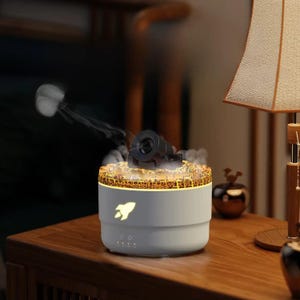 Peut inclure: Un diffuseur d'aromathérapie blanc et gris avec un design de fusée. Le diffuseur a une caractéristique noire en forme de canon sur le dessus, émettant de la vapeur. La base a un logo de fusée et des accents lumineux. Il est posé sur une surface en bois.