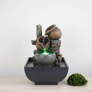 Peut inclure: Fontaine d'intérieur décorative avec un design à plusieurs niveaux. L'eau coule de deux pots en céramique marron dans un bol en pierre, éclairé par une lumière verte. La fontaine est dans un bassin noir, avec une petite plante verte à côté.